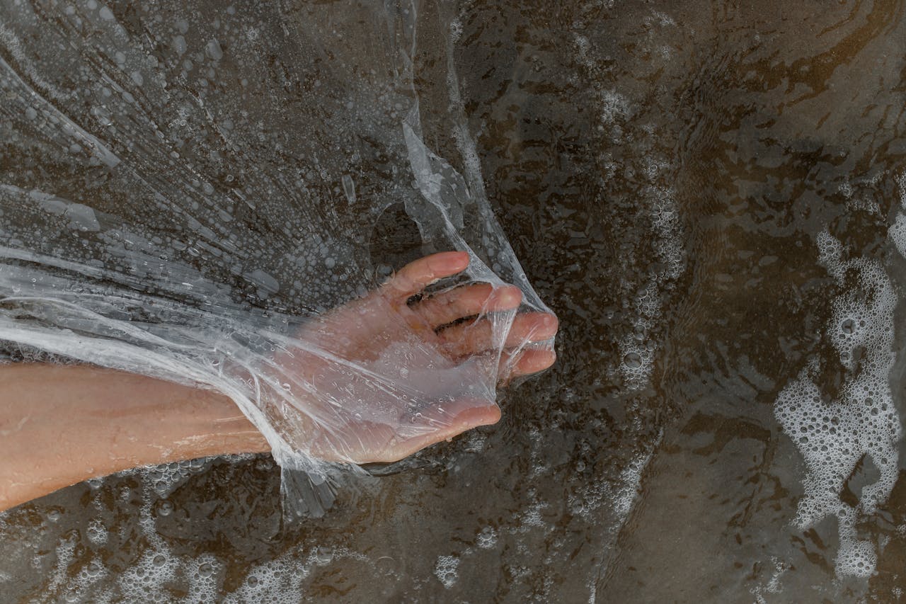 A hand holds a plastic sheet amidst seawater foam on a sandy beach, highlighting pollution.