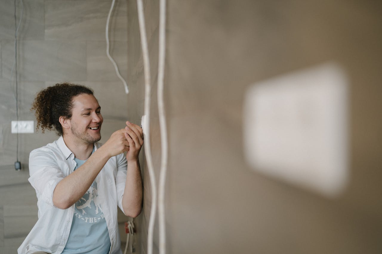 An electrician with curly hair adjusts an outlet indoors, smiling as he works.